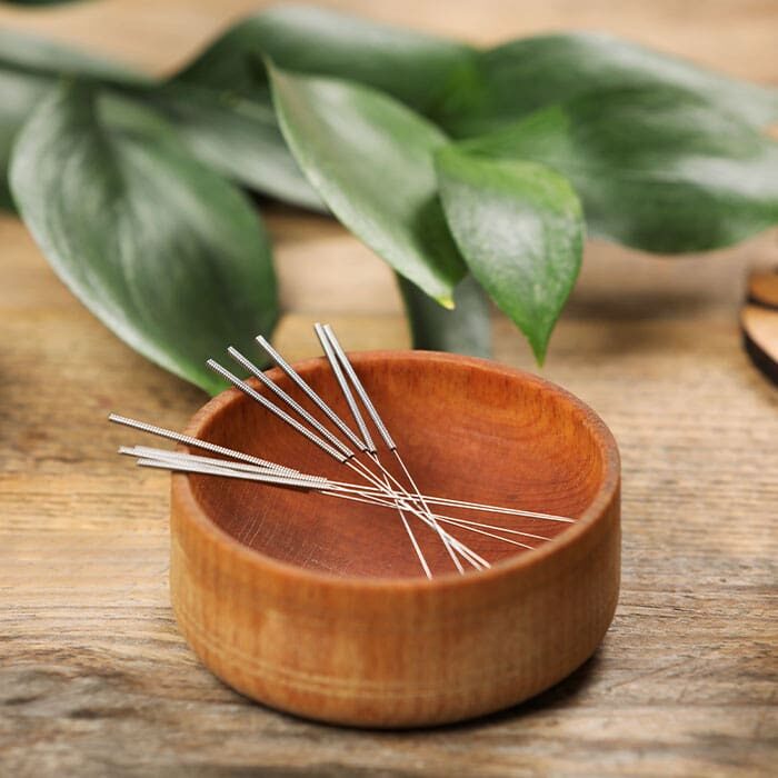 acupuncture needles sitting in shallow wooden bowl on wooden table with leaves from plant in background